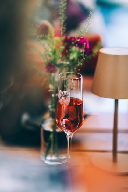 Ein Glas Rosé-Sekt auf dem Tisch, daneben Wiesenblumen und eine Tischleuchte