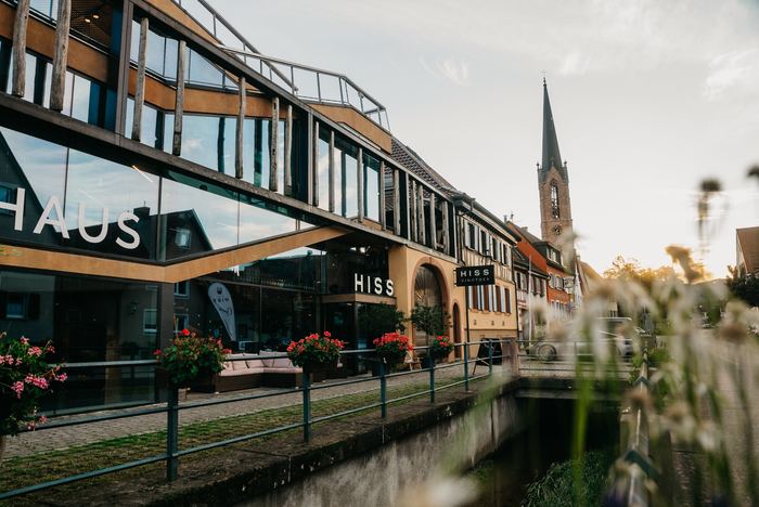 Das Weinhaus Hiss von der Straße aus mit dem Schriftzug HISS über dem Eingang, dahinter der Kirchturm von Eichstetten, im Vordergrund ein Wasserlauf mit Geranien am Geländer
